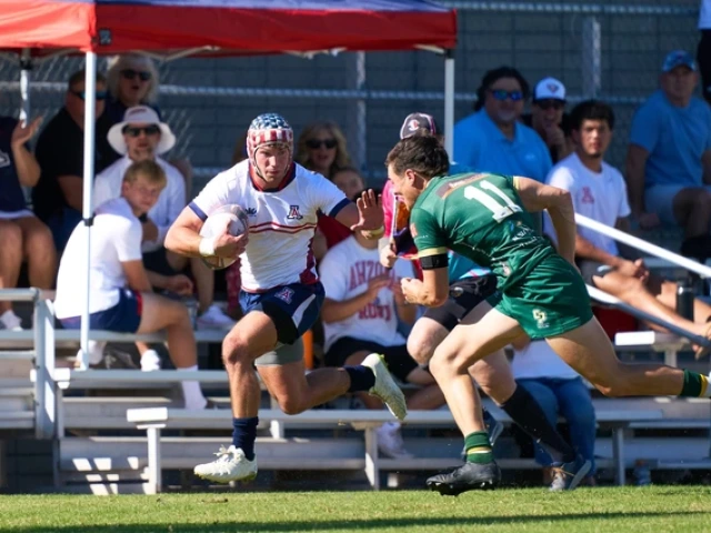 A rugby match with two male players in action. The player on the left, in a white jersey with blue and red accents, holds a rugby ball and is sprinting forward. He wears a protective headband with a star pattern. The player on the right, wearing a green jersey marked with the number 11, is approaching to tackle. Both players are on a grassy field with sunlight casting shadows.