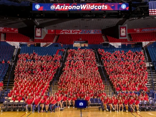 Hundreds of University of Arizona student-athletes wearing red shirts sit together in the shape of a large block “A” inside McKale Memorial Center during the Arizona Athletics welcome back BBQ. An American flag hangs above, and a scoreboard displays the @ArizonaWildcats handle.