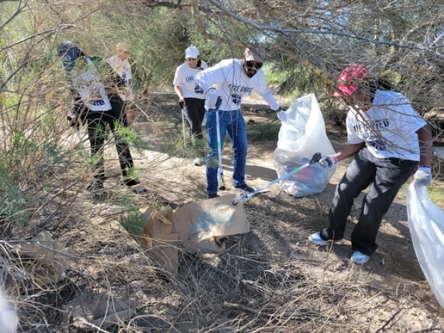 University of Arizona volunteers wearing “Live United” shirts pick up trash and debris in a wooded area using grabbers and large plastic bags during Eller Make a Difference Day.