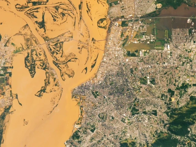 Satellite image showing a dense urban cityscape bordered by a large, muddy river with winding channels and sediment-rich water. The grid-like streets and clusters of buildings contrast with patches of green spaces, while bridges connect the city across the river.