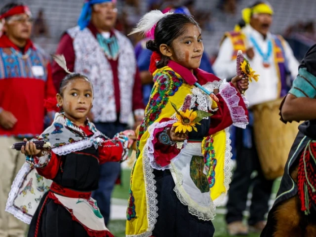 Two young children in traditional Hopi regalia perform a dance on the field, holding corn and flowers, during the Hopi Recognition Football Game at Arizona Stadium, with adult participants standing behind them.