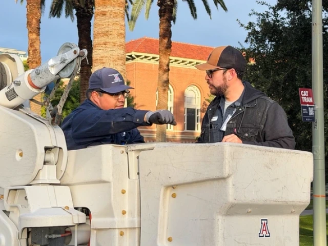 Two University of Arizona Facilities Management workers talk beside a bucket truck on campus. One worker sits inside the raised bucket, wearing gloves and a U of A–branded cap, while the other stands beside the truck, listening. Old Main is visible in the background along with palm trees.
