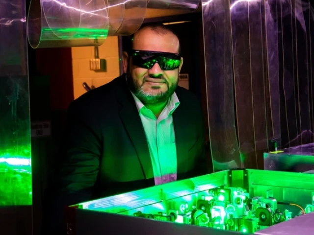 A man wearing safety glasses and a suit observes a green-lit optical experiment with lasers and mirrors in a laboratory setting.