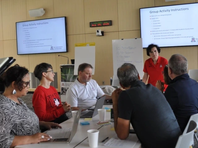  A group of six people sit around a table in a meeting room at the University of Arizona, listening to a woman in a red U of A polo shirt who is speaking to them. Laptops, papers, and coffee cups are on the table, and two screens behind them display “Group Activity Instructions.”