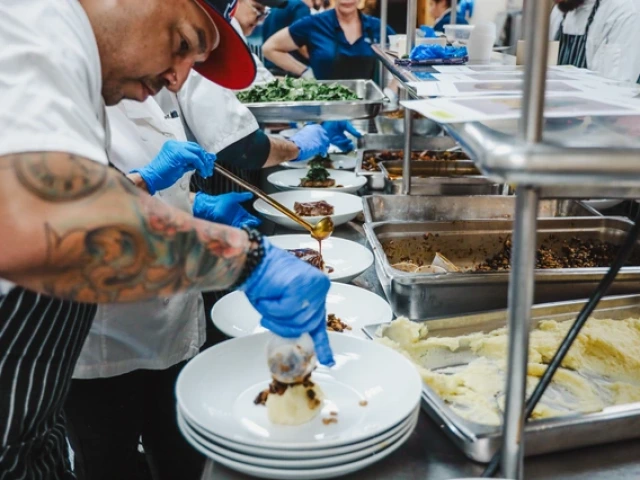 Chef in a red cap and tattooed arms plates dishes in a busy kitchen. Others assist with food preparation. Blue gloves worn, food on trays nearby.