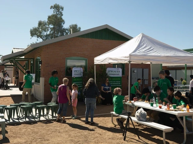 People gather outside the newly renovated Sprouts House during a sunny daytime event. A white tent shades a table where participants in green shirts sit and eat, while others talk nearby. The restored brick building with green trim stands in the background with banners showing the Sprouts Foundation’s mission.