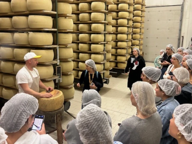 Students wearing hairnets gather around a worker in a cheese factory, listening as he explains the process in front of large wheels of cheese stacked high on shelves.