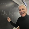 A professor wearing a black shirt writes on a chalkboard